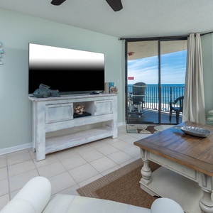 The living room features a large TV, a wooden coffee table, and a cozy chair, with a beautiful view of the beach beyond