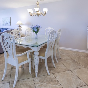 Elegant dining area features a glass-top table surrounded by white chairs, complemented by soft lighting and coastal decor