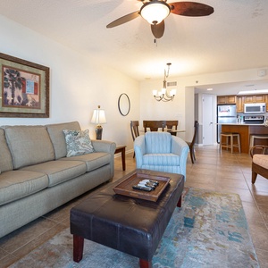 Cozy living area features a light gray sofa with tropical pillows, a dark ottoman, and a dining space leading to the kitchen