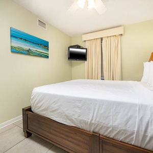 A cozy guest bedroom features a queen bed, a wall-mounted TV, and soft green walls accented by a colorful beach painting