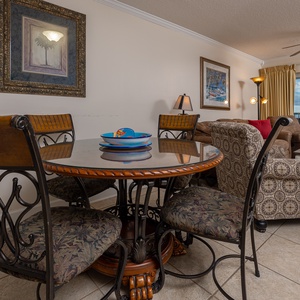 The living area features a round dining table, plush seating, and a stunning beachfront view through large sliding doors