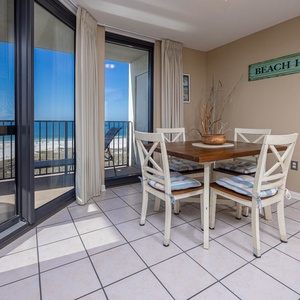 Dining area features a wooden table with cushioned chairs, offering a view of the beach through large glass doors