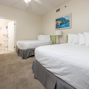 Bright guest bedroom featuring two double beds with plush bedding, a bedside lamp, and a doorway leading to a bathroom