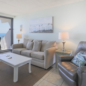 The living room features comfortable seating, a stylish coffee table, and stunning beachfront views through large windows