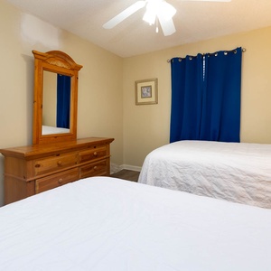 Bright guest bedroom featuring two double beds, a wooden dresser with a mirror, and blue curtains adding a pop of color