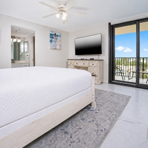 Bright master suite featuring a plush king bed, a large TV, and a sliding door leading to a balcony with views of the area