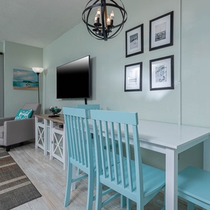 This inviting living area features a large TV, light blue dining chairs, and a stunning beachfront view from the balcony