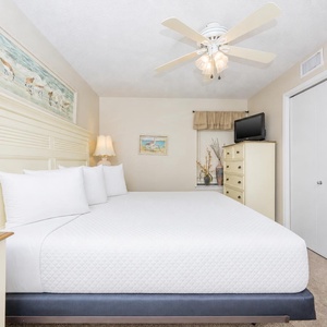 Bright master suite bedroom featuring a king bed, soft linens, a ceiling fan, and a TV with coastal art enhancing the vibe