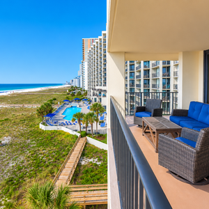 Spacious balcony features comfortable seating with vibrant blue cushions, offering a view of the beach and nearby pool area