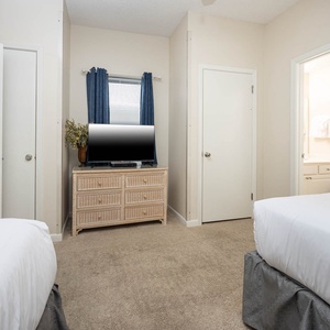 A cozy guest bedroom features two double beds, a TV on a wicker dresser, and a doorway leading to an en-suite bathroom