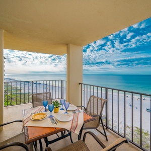 A dining table set for six overlooks a tranquil beachfront, with blue skies and gentle waves visible in the distance