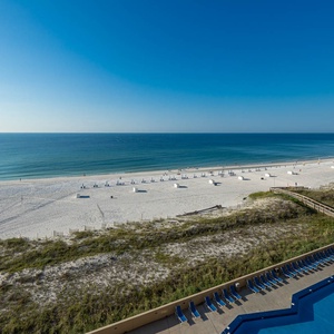 Expansive view of the beach with white sand and gentle waves, showcasing lounge chairs along the shore and the pool area