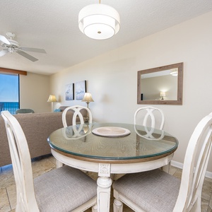 A round glass-top dining table with white chairs sits in a bright space, offering a view of the beach from the balcony