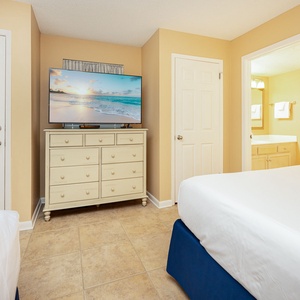 Cozy guest bedroom featuring a large TV, two beds, dresser, and a view into the bathroom with a bright, inviting atmosphere
