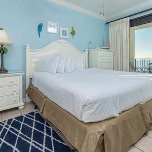 Cozy master bedroom featuring a king bed, light blue walls, and access to a balcony with a view of the beach