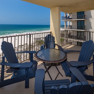 A spacious balcony features vibrant blue chairs and a table, offering a serene view of the beach and surrounding greenery