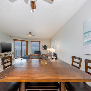 Dining area features a rustic wooden table with chairs, leading to a cozy living space with a beach view