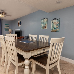 Bright dining area with a rustic table, white chairs, and beach-themed decor, offering a view of the tranquil coastline