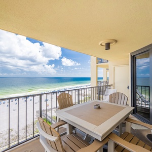 The balcony features a dining table and chairs with a breathtaking view of the beach and vibrant blue waters