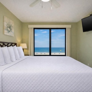 This bedroom features a plush king bed, soft white linens, and a stunning beachfront view through large windows