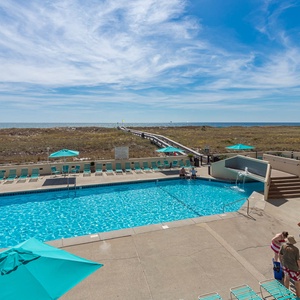 Sparkling pool area features lounge chairs and colorful umbrellas, with a scenic view leading to the beach and boardwalk