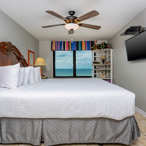 The master bedroom features a plush king bed, a mounted TV, and a stunning beachfront view from the large window