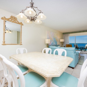 This dining area features a light stone table and white chairs, with a stunning beachfront view visible through large windows