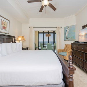 Master bedroom featuring a plush king bed and a balcony view with dining area and coastal artwork