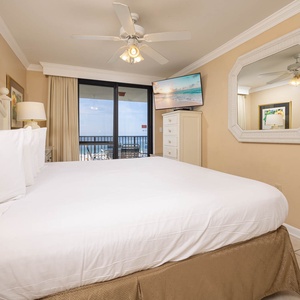 Cozy master bedroom featuring a king bed, a TV, and a balcony with views of the beach, complemented by soft, neutral decor