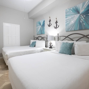 Bright guest bedroom featuring two queen beds with decorative pillows, coastal artwork, and a stylish dresser