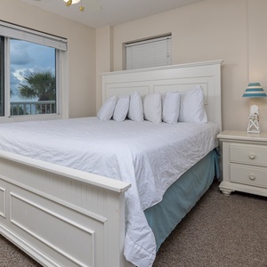 The master bedroom features a plush white bed, colorful fish art, and a stunning view of palm trees and the beach