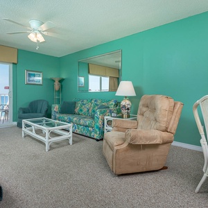 The living area features vibrant teal walls, comfortable seating, and a stunning beachfront view through sliding glass doors