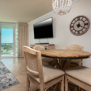 This inviting dining area features a round wooden table, comfortable chairs, and a large TV, with a lovely view beyond