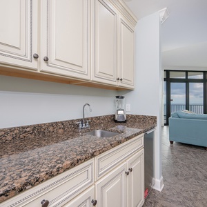 Kitchen wet bar area with granite countertops, a blender, ice-maker, and a view of the cozy living area and expansive windows