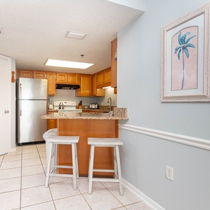 The fully-equipped kitchen features warm wood cabinetry, a breakfast bar with two stools, and a coastal-themed wall art