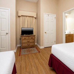 Two double beds with white linens are set against a warm-toned room featuring a TV on a wicker dresser and en-suite bathroom