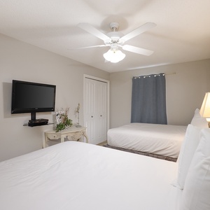 This guest bedroom features two double beds, a wall-mounted TV, and a cozy bedside lamp beside a decorative table