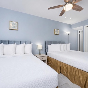 This guest bedroom features two double beds with plush white bedding, a stylish ceiling fan, and soft blue walls