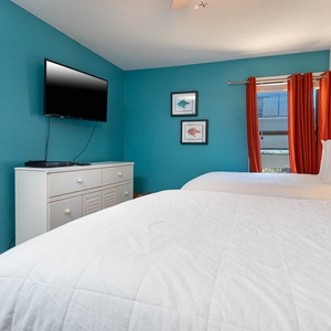 This guest bedroom features two comfortable full beds, a wall-mounted TV, and vibrant orange curtains against a teal backdrop