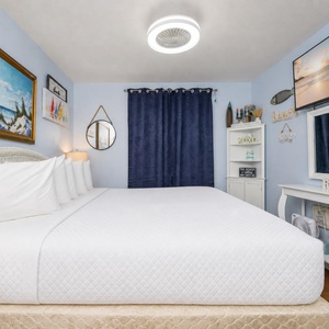 The bedroom features a cozy white bed adorned with plush pillows, beach-themed artwork, and soft lighting for a relaxing stay