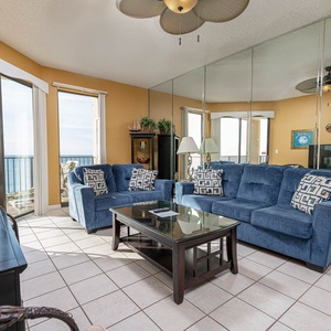 Cozy living room featuring blue sofas with decorative pillows, a glass coffee table, and large windows showcasing the beach