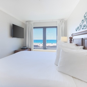 The master bedroom features a plush bed and a stunning beachfront view through large windows, perfect for relaxation