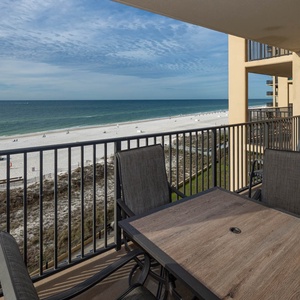 A spacious balcony features a wooden table and chairs, offering a serene view of the beach and tranquil blue waters