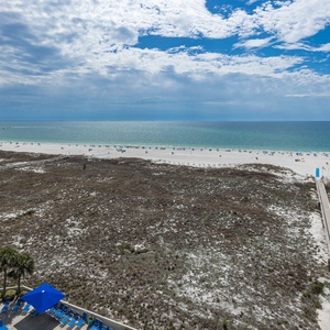 Expansive view of the beach with white sand, turquoise gulf waters, and lounge chairs lining the shore under a bright sky