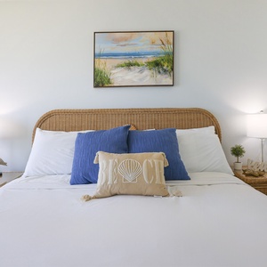 The bedroom features a cozy rattan headboard, soft blue accents, and beach-themed decor for a relaxing getaway