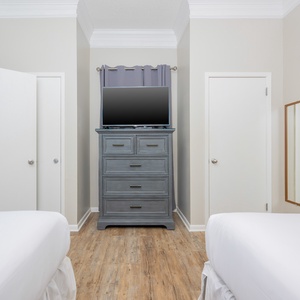 Two double beds with white linens flank a gray dresser holding a TV, leading to a bathroom with a mirror and light decor