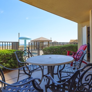 A patio area features a round table surrounded by black metal chairs, with a glimpse of the beach and pool area