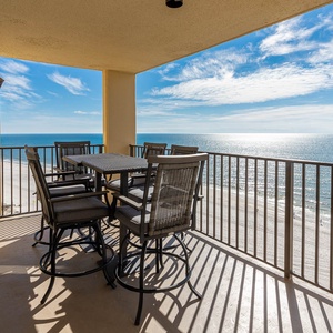 A spacious balcony features a dining set with high-backed chairs, offering a serene view of the shimmering beachfront