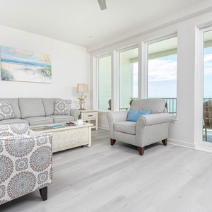 The living area features comfortable seating, a coastal painting, and stunning beachfront views through large windows