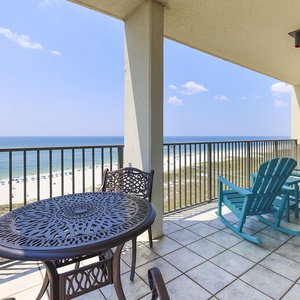Balcony features a round table and turquoise rocking chairs, offering a serene view of the beach and gentle waves below